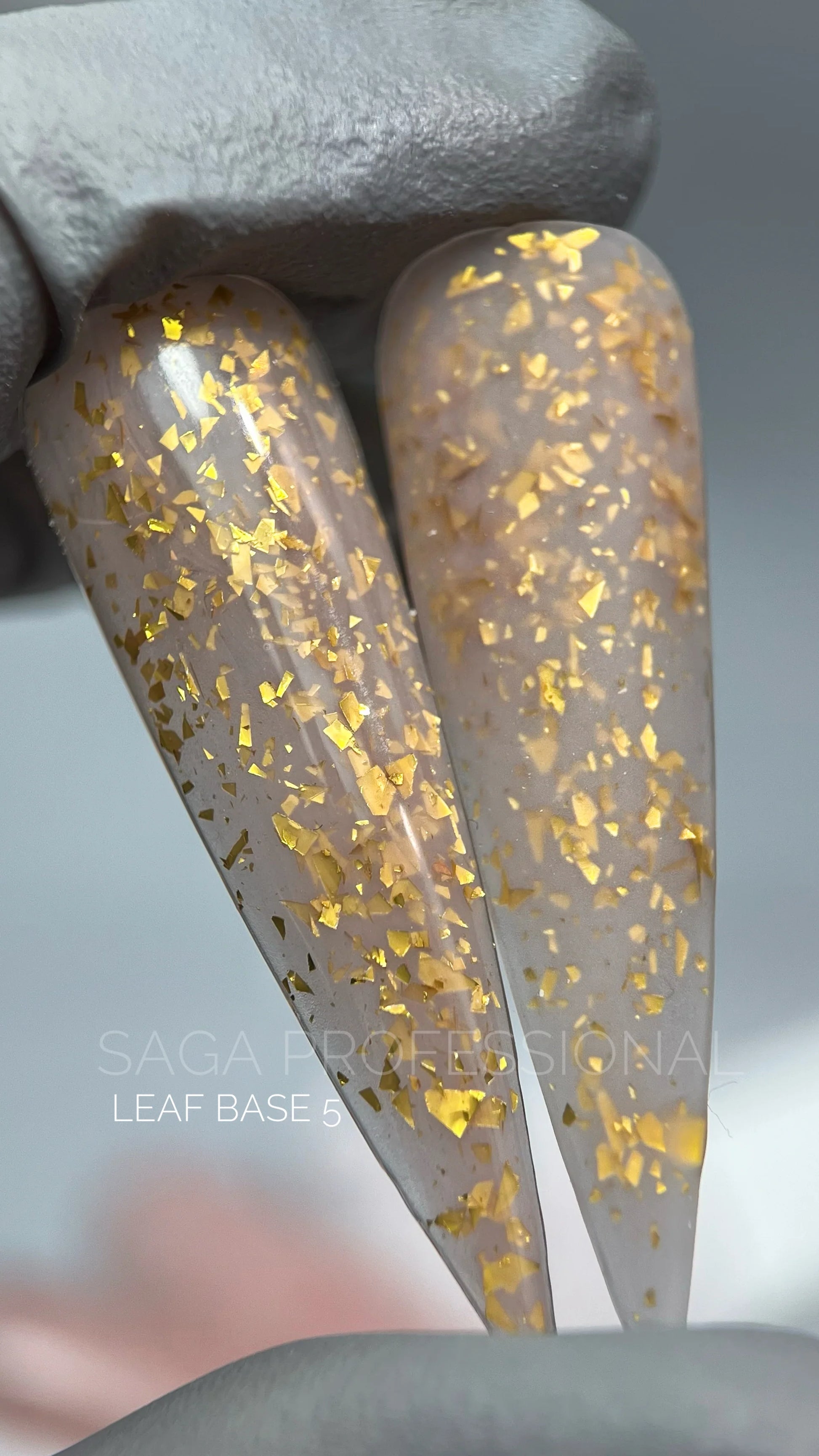 alt= Close-up of two fingernail-shaped samples with pale nail polish and gold foil flakes, held by a gloved hand. Text reads "Saga Professional Leaf Base 5" at the bottom.